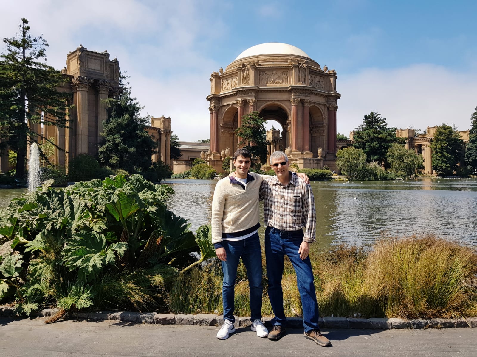 Two visitors standing in front of the Palace of Fine Arts dome and lagoon with ornamental plants in foreground