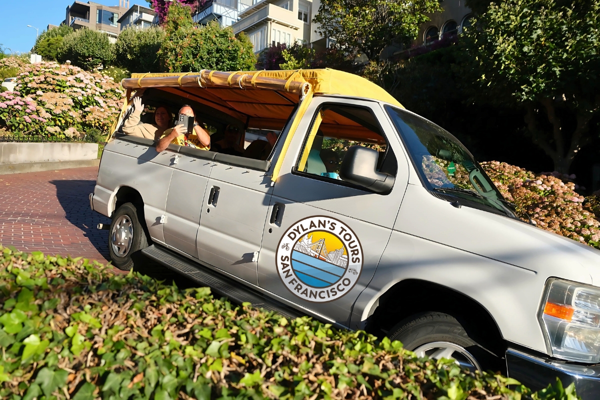 Gray Dylan's Tours van with yellow canopy driving through flowering hydrangea gardens on Lombard Street's brick roadway