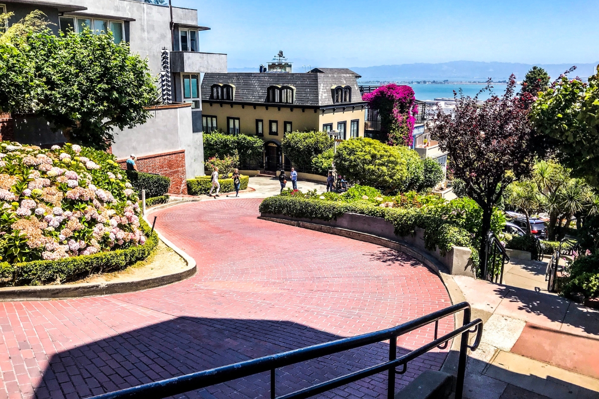 The iconic red brick switchbacks of Lombard Street in San Francisco with lush gardens, Victorian homes, and views of the bay, showcasing the street from both street level and aerial perspectives.