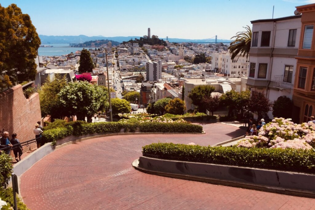 Aerial view of Lombard Street's iconic red brick switchbacks with flowering gardens, Victorian houses, and panoramic San Francisco cityscape with Bay in background